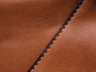 The Fur Cache Close-up of brown leather with visible grain texture and a diagonal seam stitched with white thread, bordered by a thin black line. The lighting highlights the smooth and slightly glossy surface. The best place to buy and sell furs, leather, pelts, leather products and more.