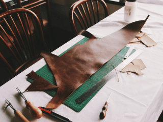 The Fur Cache A piece of brown leather, cutting tools, a ruler, and templates are arranged on a white table, suggesting leathercraft work in progress. Wooden chairs are in the background. The best place to buy and sell furs, leather, pelts, leather products and more.